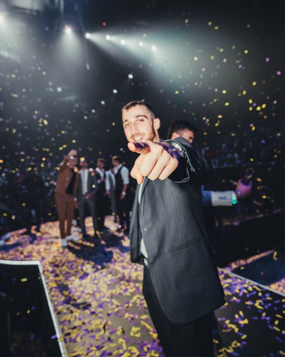 Un relatore sul palco indica il pubblico tra i coriandoli luminosi durante un grande evento