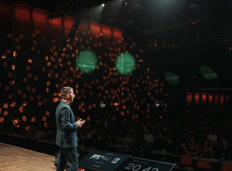 Un relatore da solo sul palco di una grande sala, con il pubblico in penombra