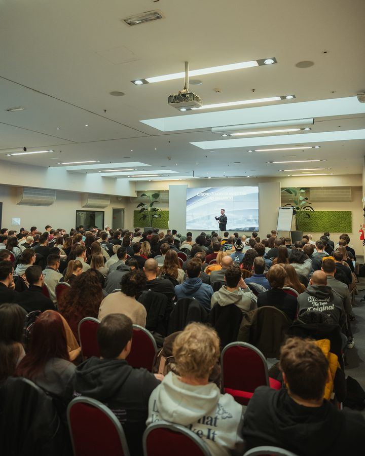 Una sala piena di persone sedute ascolta una presentazione, con il relatore in fondo