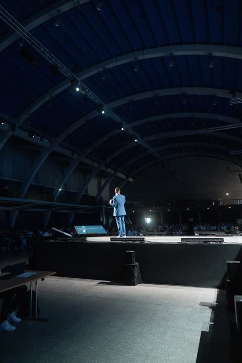 Un relatore in piedi sul palco di una grande arena buia, con le luci puntate sulla sala
